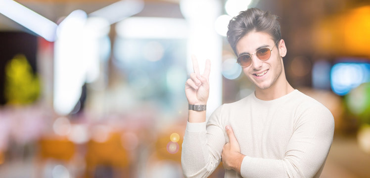 Young handsome man wearing sunglasses over isolated background smiling with happy face winking at the camera doing victory sign. Number two.
