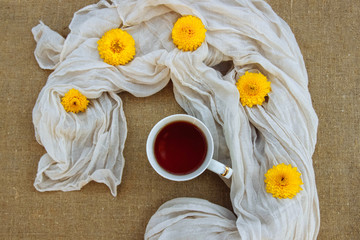 White mug with tea on the table and linen tablecloth with light scarf and yellow chrysanthemum flowers top view
