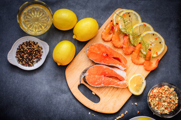 On a gray tabletop runs a wooden board on which salmon raw shrimp lemon slices pepper oil seasoning.