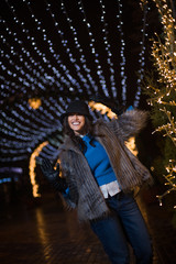 Pretty dark haired girl wearing a fur coat, blue jeans, blue top and a black hat, smiling, posing with snowflakes Christmas lights outdoor at night time.