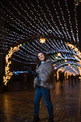 Obraz na płótnie Canvas Pretty dark haired girl wearing a fur coat, blue jeans, blue top and a black hat, smiling, posing with snowflakes Christmas lights outdoor at night time.