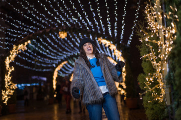 Pretty dark haired girl wearing a fur coat, blue jeans, blue top and a black hat, smiling, posing with snowflakes Christmas lights outdoor at night time.