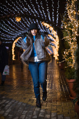 Pretty dark haired girl wearing a fur coat, blue jeans, blue top and a black hat, smiling, posing with snowflakes Christmas lights outdoor at night time.