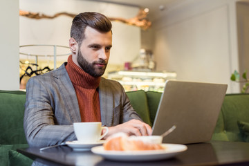 focused handsome businessman in formal wear sitting at table and typing on laptop in restaurant