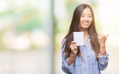 Young asian woman holding notebook over isolated background pointing and showing with thumb up to the side with happy face smiling