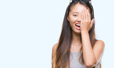 Young asian woman wearing sunglasses over isolated background covering one eye with hand with confident smile on face and surprise emotion.