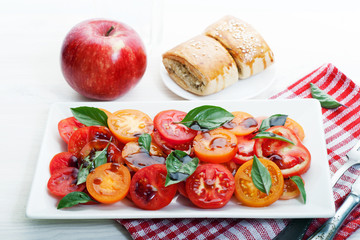Light organic salad with tomatoes, balsamic vinegar, olive oil, salt and basil leaves in a rectangular dish on a white wooden board. Next to gluten-free buns with sesame, apple and a glass of water. 