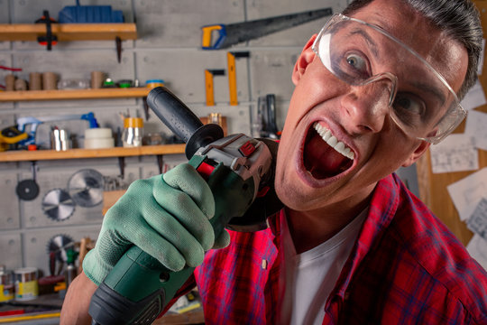 Crazy aggressive batty mad fun young man in plaid shirt, safety eye glasses on for protection, gloves with circular saw in the carpentry workshop