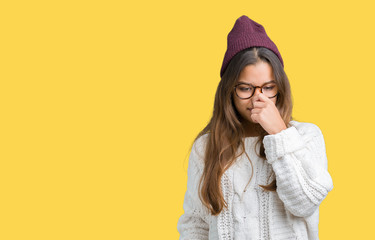 Young beautiful brunette hipster woman wearing glasses and winter hat over isolated background smelling something stinky and disgusting, intolerable smell, holding breath with fingers on nose