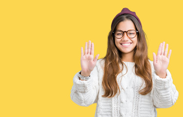 Young beautiful brunette hipster woman wearing glasses and winter hat over isolated background showing and pointing up with fingers number ten while smiling confident and happy.