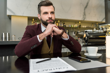 pensive businessman sitting at table with contract, coffee and smartphone while looking away