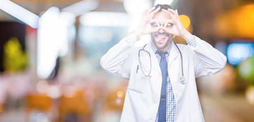 Handsome young doctor man over isolated background doing ok gesture like binoculars sticking tongue out, eyes looking through fingers. Crazy expression.