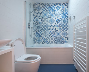 Modern bathroom with bath, toilet, niche in wall and basin unit, blue rubber floor and blue and white patchwork tiles above the bath. 