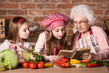 Granny with two granddaughters reading recipe from cooking book and cooking salad