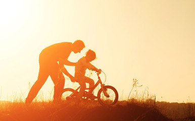 Silhouette of father teaching little daughter to ride a bike at meadow during sunset. Caring father teaches her daughter to ride bicycle