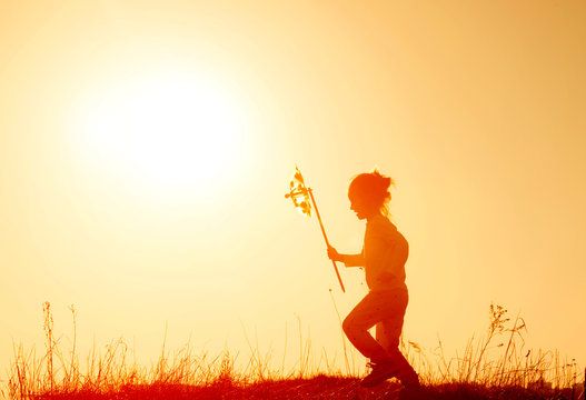 Little Girl Running On The Meadow At Sunset With Windmill In Her Hands. Silhouette Of Child Girl Holding Wind Toy