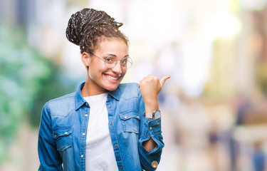 Young braided hair african american girl wearing glasses over isolated background smiling with happy face looking and pointing to the side with thumb up.