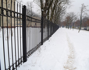 Metal long black fence near the road in the winter city.