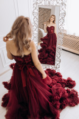 Beautiful blonde woman in luxury red burgundi dress poses before a mirror in a white room