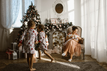 Obraz na płótnie Canvas Two little sisters in pajamas having fun New Year's tree with gifts in the light cozy room and their mother sits in the armchair with little baby next to the fireplace