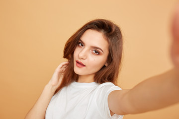 Fototapeta premium Brunette girl with long hair dressed in white t-shirt makes a selfie on the beige background in the studio