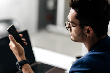 Professional architect in glasses dressed in blue checkered uses the phone near the desk with laptop in the office