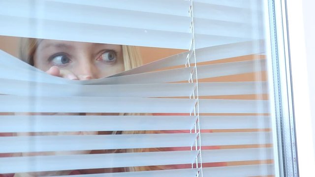 Young woman spying, peeking through the blinds in her home