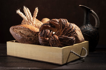 Several loafs of bread in white rustic wooden box with ears of wheat in front of dark background.