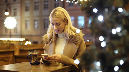 Woman using smartphone on christmas market. Girl enjoying winter holiday season. Concept of  social networking, communicating, using app. Blurred Christmas lights on background.