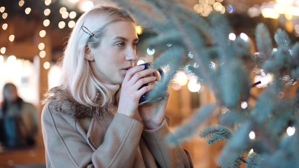 Young blonde girl drinking hot tea at christmas market in a city. Happy attractive woman in a christmas market at night. Beautiful bokeh lights background.