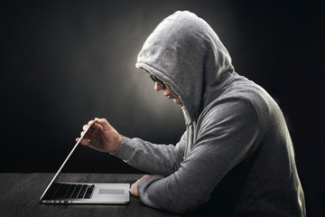 a hacker in a hood in profile sits at a laptop and starts to close it - obrazy, fototapety, plakaty