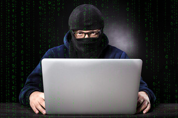 binary code on the background of a cybercriminal in a balaclava and glasses sits at a laptop