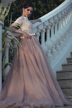 Beautiful lady with perfect make up in luxurious ballroom dress with tulle skirt and lacy top standing on stairs leaning on baluster railing. Vintage style
