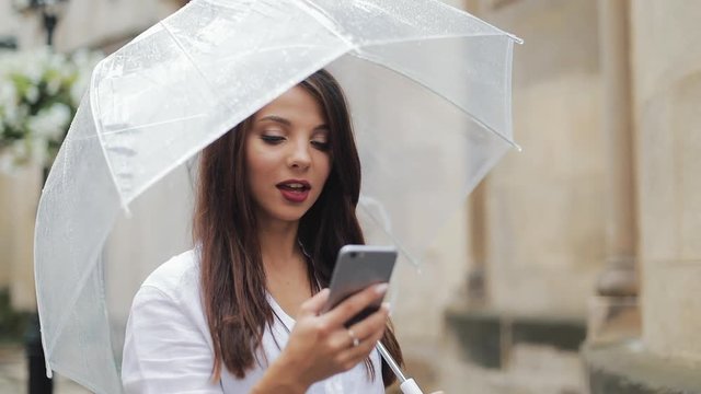 Beautiful young business woman using smartphone walking on the street in rainy weather, smiling, holding umbrella. Communication concept