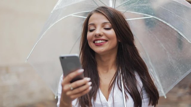 Beautiful young business woman using smartphone walking on the street in rainy weather, smiling, holding umbrella. Communication concept