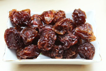 Black plum black plum dry in the plate . Prunes in a square white bowl on a white background. Prunes are the dried fruit group . Raisin in the bowl on the wood background