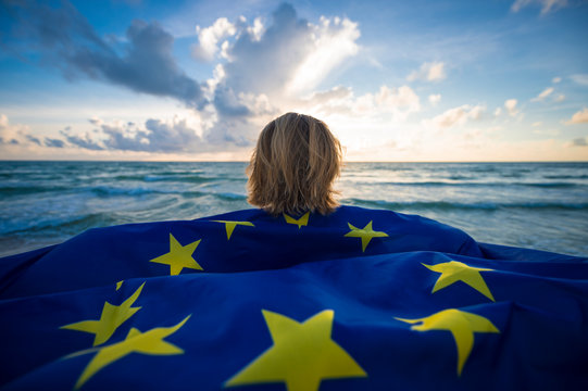 Man Holding A Fluttering Iconic EU Flag With Circle Of Stars On Beach With Stormy Turbulent Seas In The Channel At Sunrise