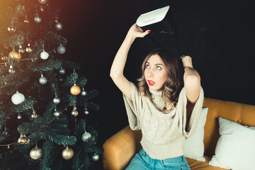 Attractive young girl holds in her hand a headset for virtual reality. Model sitting by a festive Christmas tree decorated with toys. Concept of the headset as a gift. Trend in the world of gifts