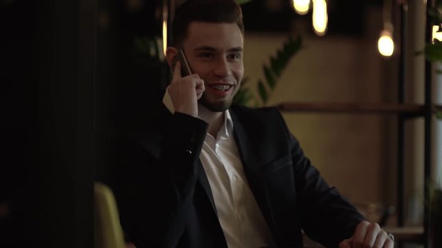 Stout handsome young man with black beard in classic suit and white shirt sitting in a cozy cafe talking on the phone, laughing, showing broad smile with braces on teeth. Having a look on his watch