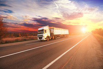 Trucks with a trailers and cars on the countryside road against sky with sunset