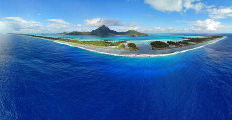 Aerial panoramic landscape view of the island of Bora Bora in French Polynesia with the Mont Otemanu mountain surrounded by a turquoise lagoon, motu atolls, reef barrier, and the South Pacific Ocean