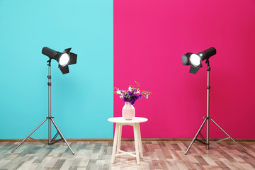 Professional lighting equipment and vase with flowers on table near wall in photo studio