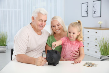 Little girl putting money into piggy bank and her grandparents at table