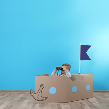 Cute little boy playing with cardboard ship near color wall. Space for text