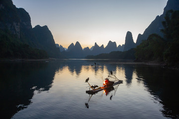 Cormorant fisherman on raft in lake in Guilin, China, with three cormorant birds. Fisherman is using a bright flame to heat teapot and light pipe.