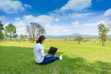 Naklejka na ściany i meble Portrait of pretty young woman sitting on green grass in park with legs crossed during summer day while using laptop