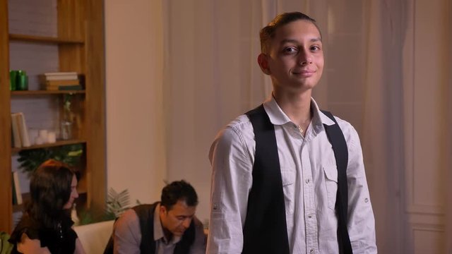 Portrait of teenage relaxed arabic boy watching directly into camera with modest smile and his family on background.