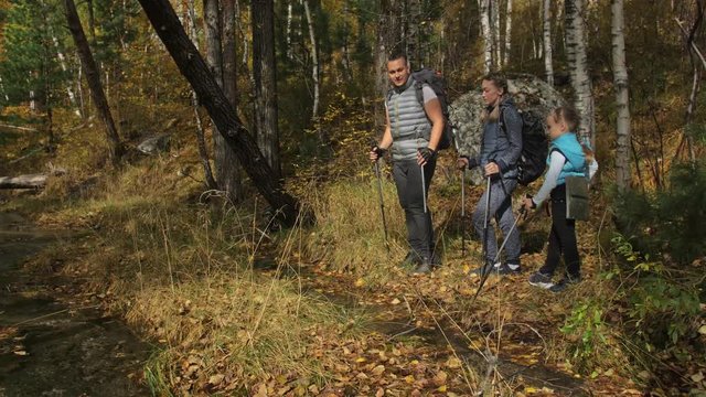 People walk near a mountain stream. Family travels. People environment by mountains, rivers, streams. Parents and kids walk using trekking poles. Man and woman have hiking backpacks, flasks, mugs. Dad