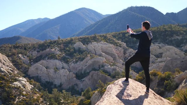 An attractive millennial man taking nature photos of an incredible landscape mountain view on a hike in the California forest.