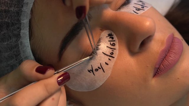 Eyelahes extension expert with tender hands which nails painted in claret polish is smoothing and streching each client's eyelash out with two special tweezers. Customer lying with under eye tape on